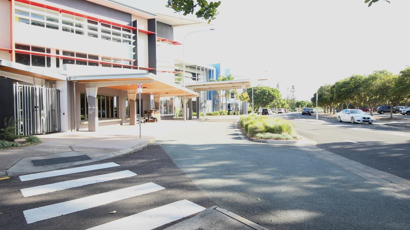 On the left, there is a modern building with red and grey accents, large windows, and a covered walkway with a beige ceiling. On the right, there are trees and parked cars along a road. In the middle, a continuation of the building extends towards the background, featuring more windows and a blue vertical stripe. The foreground includes a pedestrian crossing with white stripes, a paved walkway, and a section of asphalt road with a small landscaped area containing grass and shrubs.