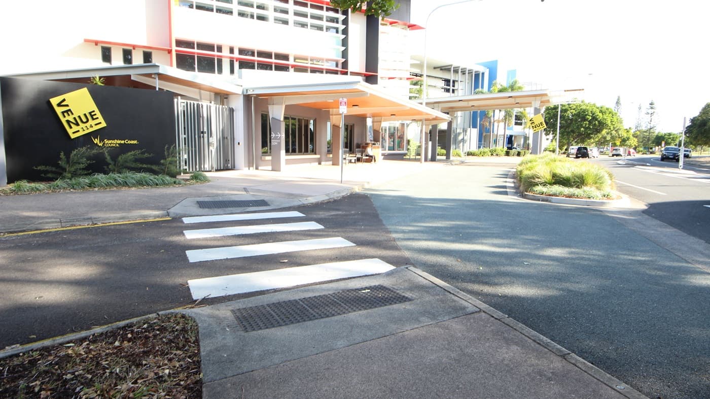 On the left, there is a building with a large yellow sign reading "VENUE" and a black wall with plants in front. On the right, there are trees and a road with parked cars. In the middle, a building with red and white accents and large windows extends towards the background, with a covered walkway featuring a beige ceiling. The foreground includes a pedestrian crossing with white stripes, a paved walkway, and a section of asphalt road with a small landscaped area containing grass and shrubs.