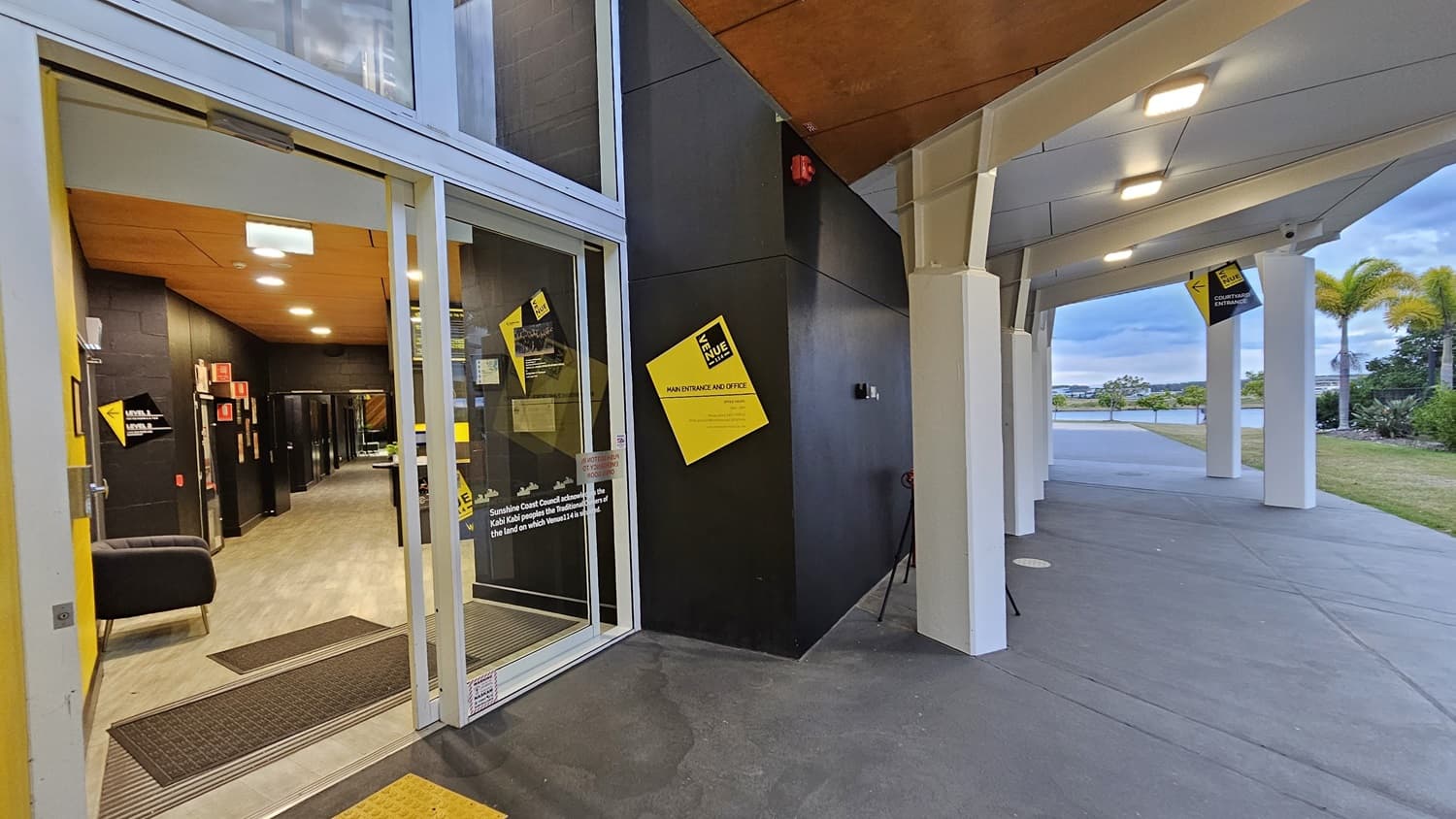 On the left, there is a glass entrance door with a yellow sign and a black sofa inside. The floor is light-coloured wood. On the right, there is a covered walkway with white pillars and a grey concrete floor. In the middle, a black wall with a yellow sign and a fire alarm. The background features a grassy area with palm trees and a body of water. The ceiling is wooden on the left and white on the right.