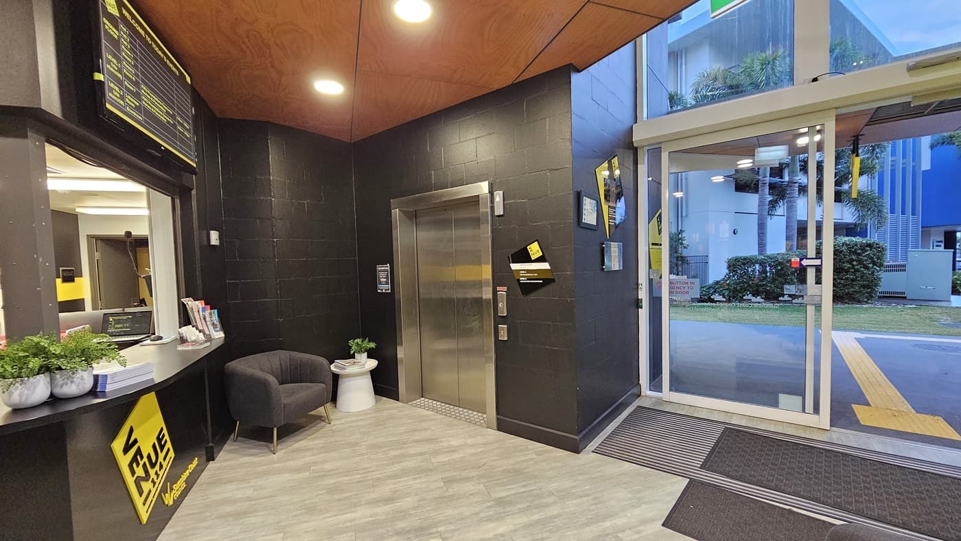 On the left, there is a reception desk with a yellow sign, a black chair, and a small white table with plants. On the right, there is a glass entrance door with a yellow sign and a view of a grassy area and buildings outside. In the middle, there is a lift with a yellow stripe and a black wall. The floor is light grey tile, and the ceiling is wooden with recessed lights.