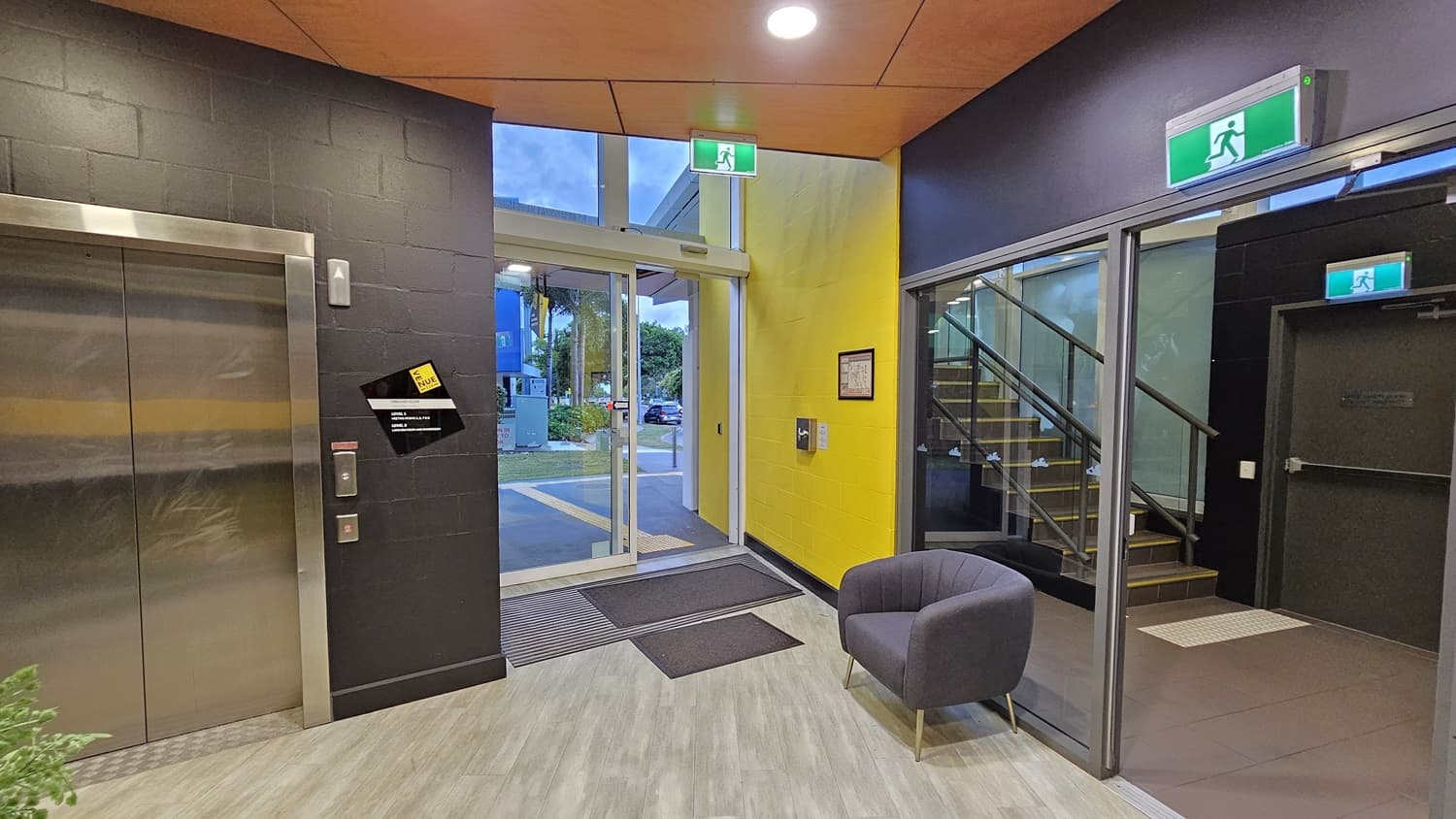 On the left, there is a lift with a black wall and a yellow stripe. On the right, there is a glass door with a green exit sign above it, leading to a staircase with a black railing. In the middle, there is a glass entrance door with a view of a car park and greenery outside. The floor is light grey wood-effect tile, and the ceiling is orange with recessed lights. A grey armchair is placed near the entrance.