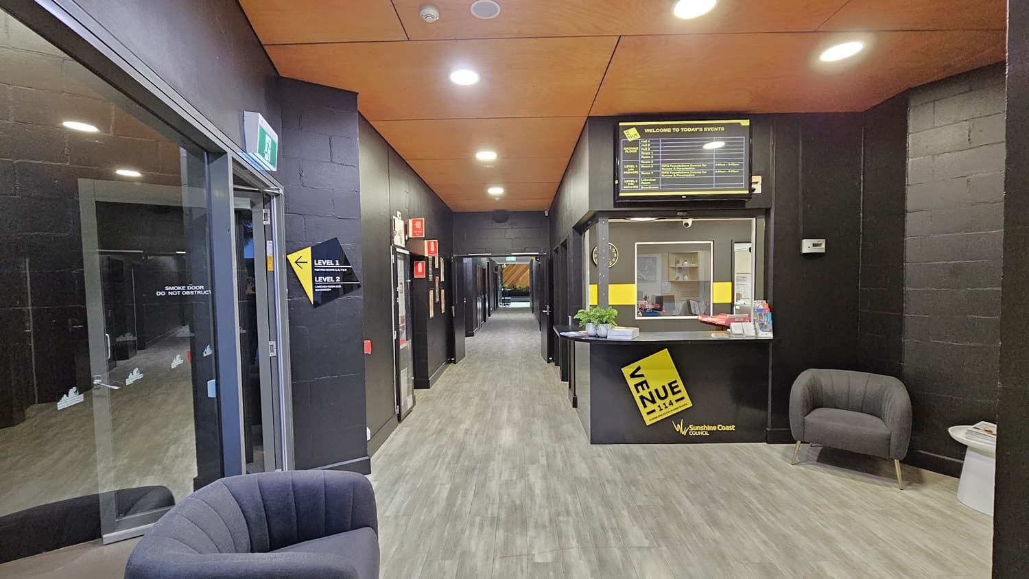 On the left, there is a glass door with a green exit sign above it, leading to a corridor with black walls. On the right, there is a reception desk with a yellow sign, a computer, and a small counter with brochures. In the middle, there is a hallway with black walls and several doors. The floor is grey wood-effect tile, and the ceiling is orange with recessed lights. Two grey armchairs are placed near the entrance.