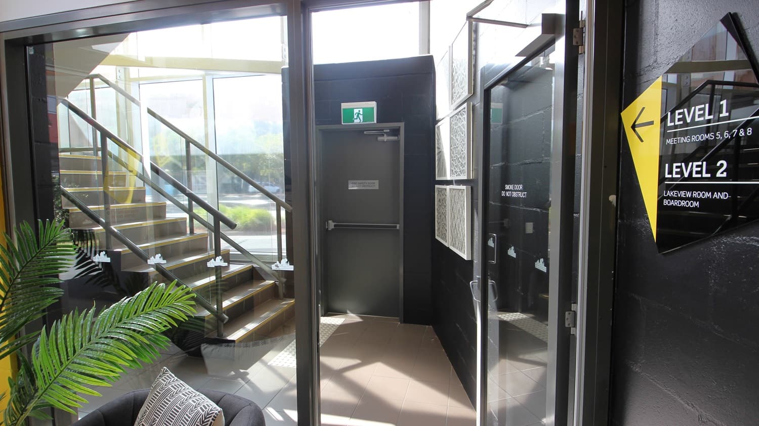 On the left, there is a staircase with black railings and wooden steps, leading to an upper level. On the right, there is a black wall with a yellow and black sign indicating "Level 1" and "Level 2". In the middle, there is a grey door with a green exit sign above it. The floor is light brown tile, and the ceiling is glass, allowing natural light to enter. A black armchair with a patterned cushion is in the foreground near a potted plant.