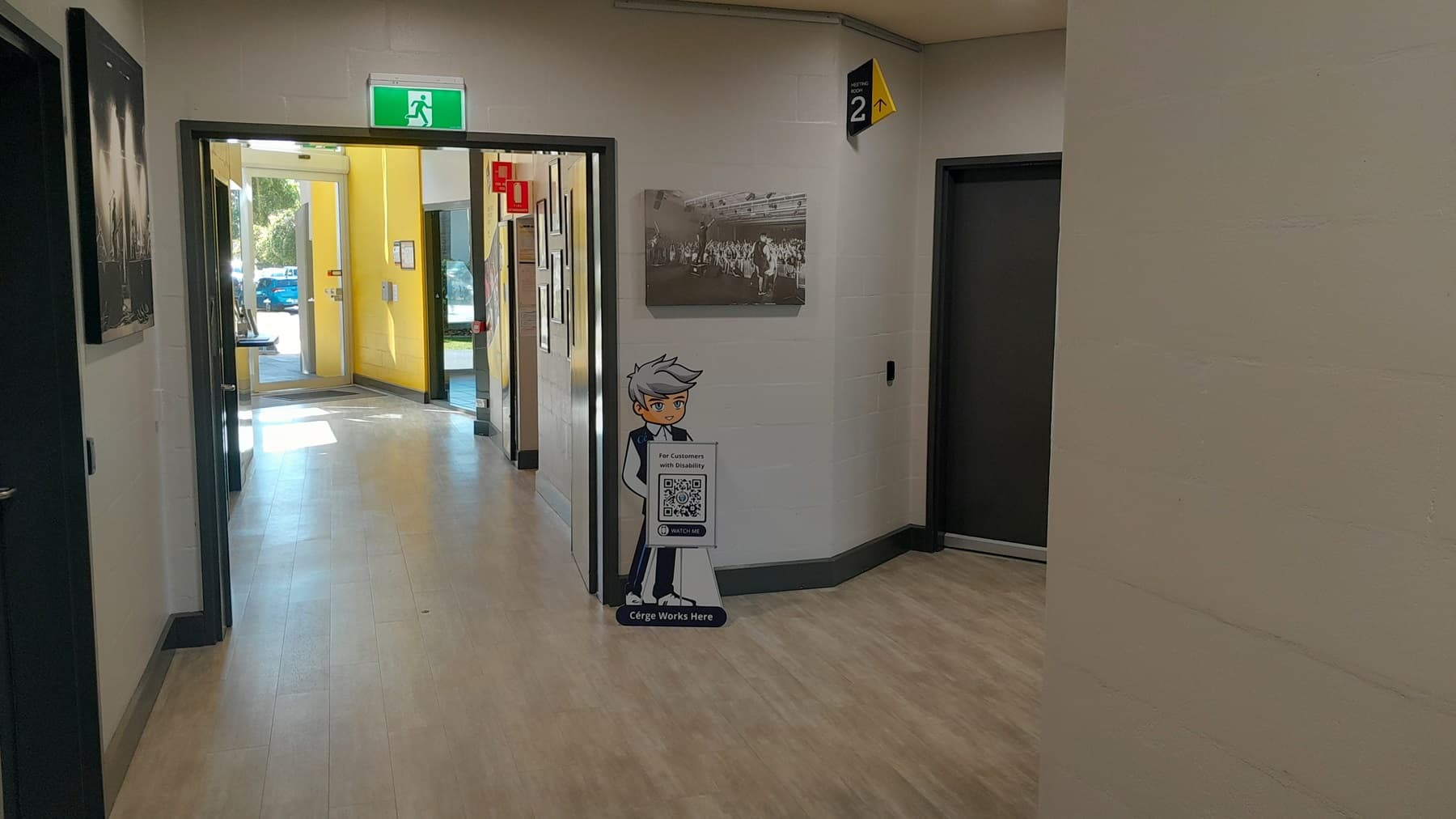On the left, there is a black and white photograph on the wall and a corridor leading to a room with yellow walls. On the right, there is a black door with a yellow and black "Level 2" sign above it. In the middle, there is a green exit sign above a glass door leading outside, with a standee of a cartoon character holding a QR code. The floor is light wood-effect tile, and the ceiling is white with recessed lights.
