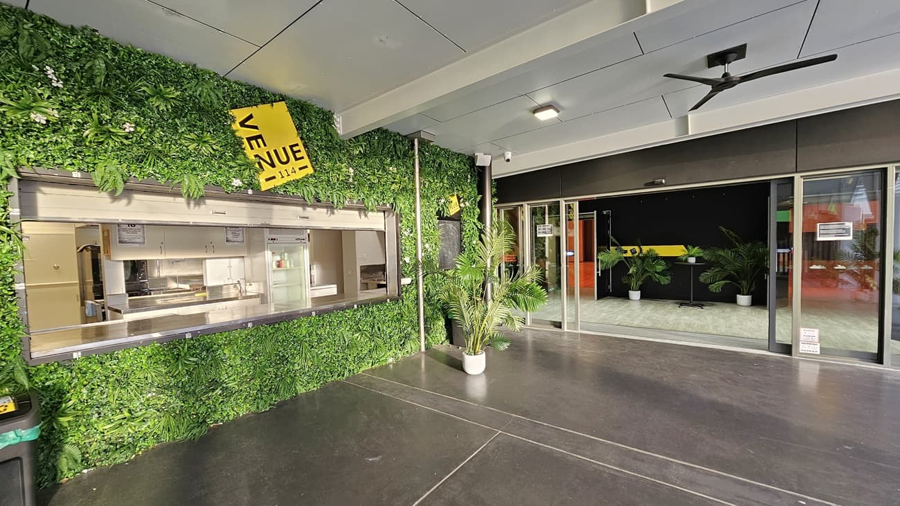 On the left, there is a service counter with a metal shutter, set within a green wall covered in artificial foliage and a yellow sign reading "Venue". On the right, there are glass sliding doors leading to an indoor area with potted plants and a black wall. In the middle, there is an open space with a dark concrete floor. The ceiling is white with recessed lights and a black ceiling fan.
