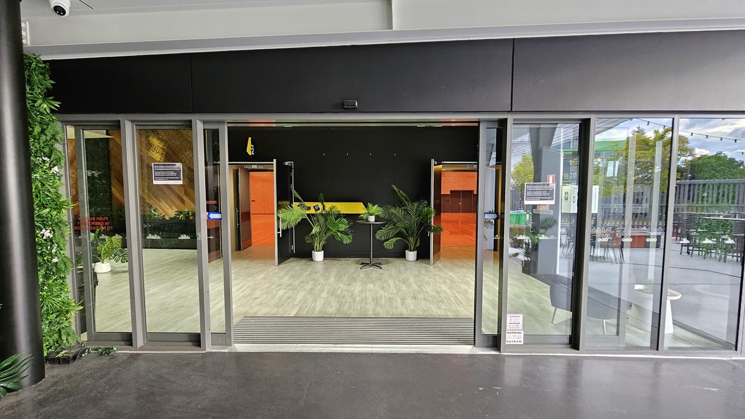 On the left, there is a green wall with artificial foliage and a black pillar. On the right, there are glass doors leading to an outdoor seating area with tables and chairs. In the middle, there is a reception area with a yellow counter, potted plants, and orange doors on either side. The floor is light grey wood-effect tile, and the ceiling is white with recessed lights.