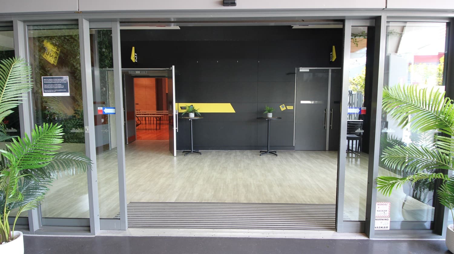 On the left, there is a green wall with artificial foliage and a small sign. On the right, there is a black wall with a door and a yellow sign above it. In the middle, there is an open entrance with glass sliding doors leading to a room with a light wood-effect tile floor. The room has a black wall with a yellow stripe, two small black tables with plants, and an open door to another room with orange walls. The ceiling is white with recessed lights.