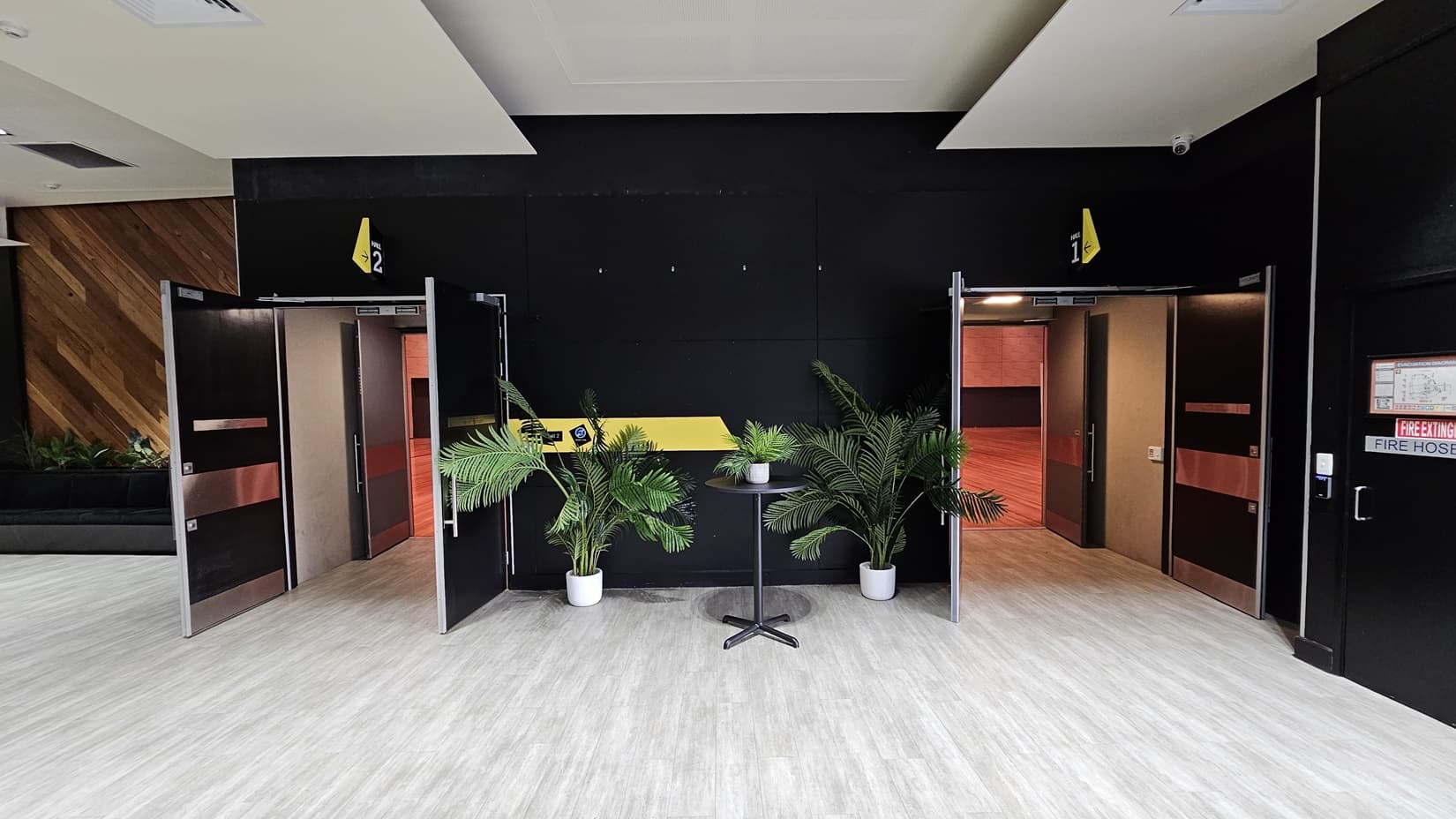 On the left, there is an open door leading to a room with red walls and wooden flooring. On the right, there is another open door leading to a similar room. In the middle, there is a black wall with a yellow stripe, two potted plants, and a small round table with a plant on it. The background features black walls with yellow signs above the doors. The floor is light grey wood, and the ceiling is white with recessed lights.