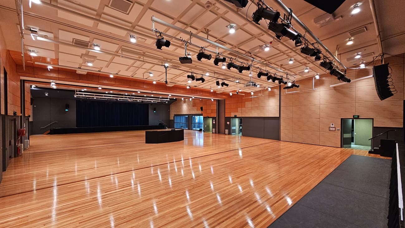 On the left, there is a stage with blue curtains and black walls, along with a fire extinguisher mounted on the wall. On the right, there is a beige wall with a green exit door. In the middle, there is a large open space with polished light wood flooring and a black equipment table. The background features beige and orange acoustic panels on the walls. The ceiling is white with a grid pattern and numerous recessed lights, along with black lighting equipment suspended from metal tracks.