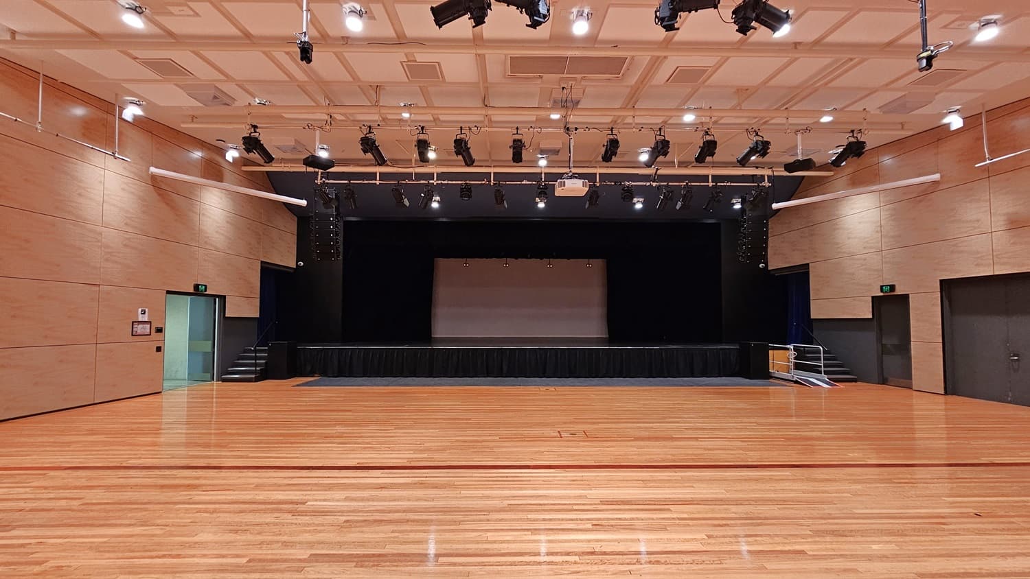 On the left, there is a beige wall with a green exit door and a fire extinguisher mounted on the wall. On the right, there is another beige wall with a dark grey door. In the middle, there is a large stage with black curtains and lighting equipment above. The background features beige walls and a black stage area. The floor is polished light wood, and the ceiling is white with a grid pattern and numerous recessed lights, along with black lighting equipment suspended from metal tracks.