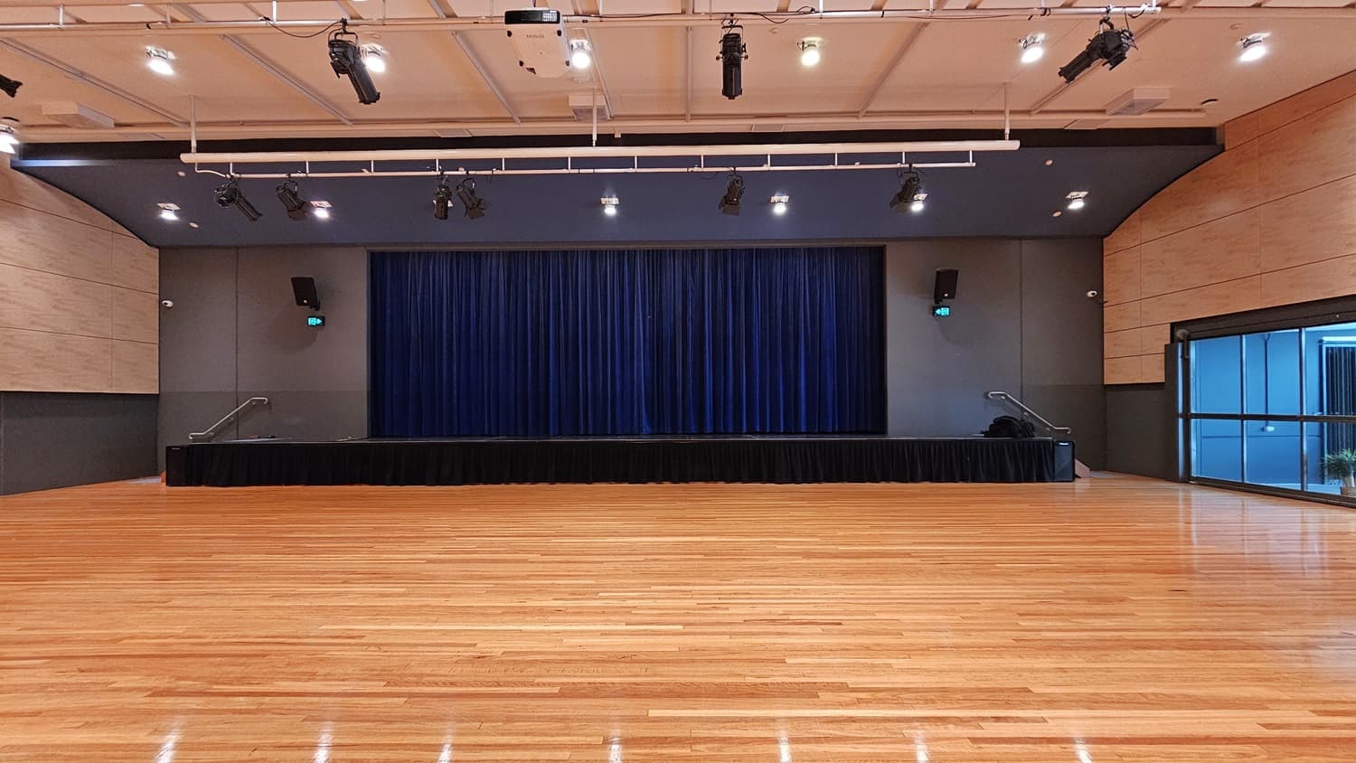 On the left is a beige wall with a black lower section and a green exit sign above a door. On the right, there is a similar beige wall with a black lower section and a green exit sign above another door. In the middle, there is a stage with blue curtains and black skirting. The background features the stage area with blue curtains. The floor is polished light wood, and the ceiling is white with a grid pattern and recessed lights, along with black lighting equipment suspended from metal tracks.