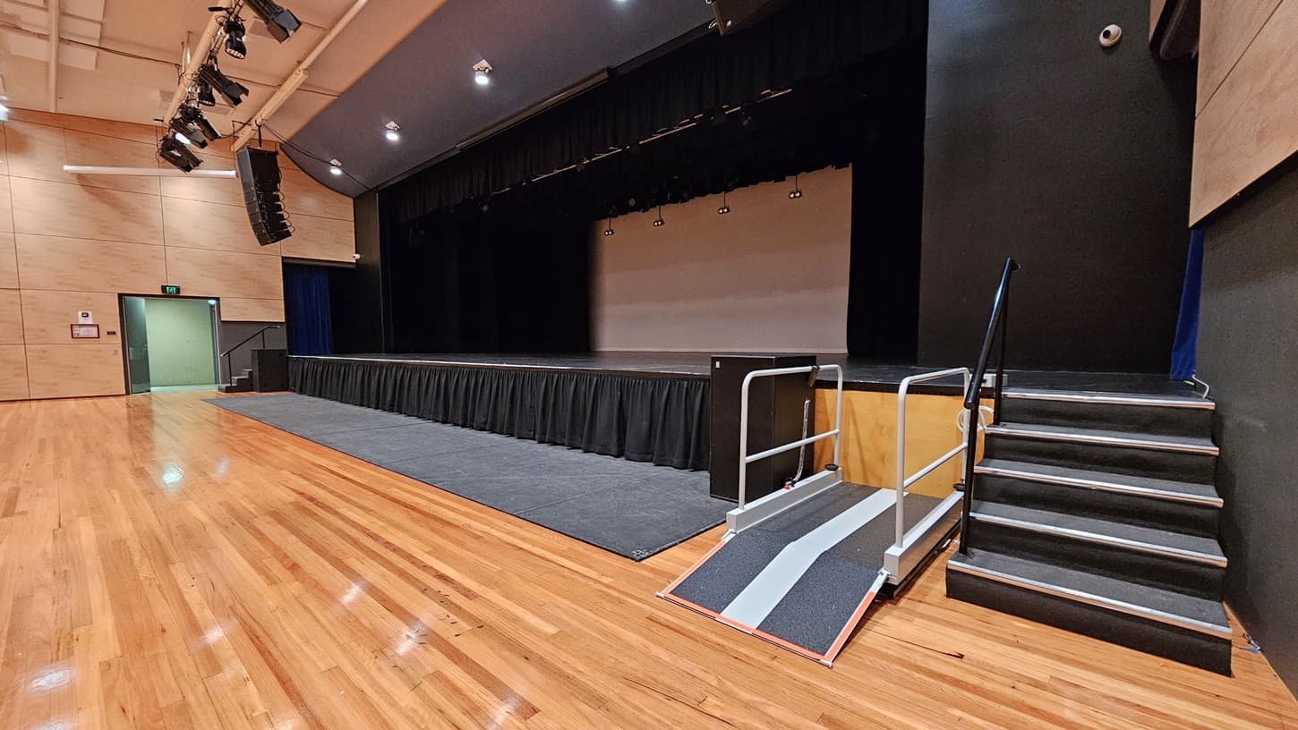On the left, there is a beige wall with a green exit door and a fire alarm. On the right, there is a black wall with a set of stairs leading up to the stage. In the middle, there is a large stage with black curtains and a grey ramp with handrails. The background features the stage area with black curtains and lighting equipment above. The floor is polished light wood, and the ceiling is white with recessed lights and black speakers.