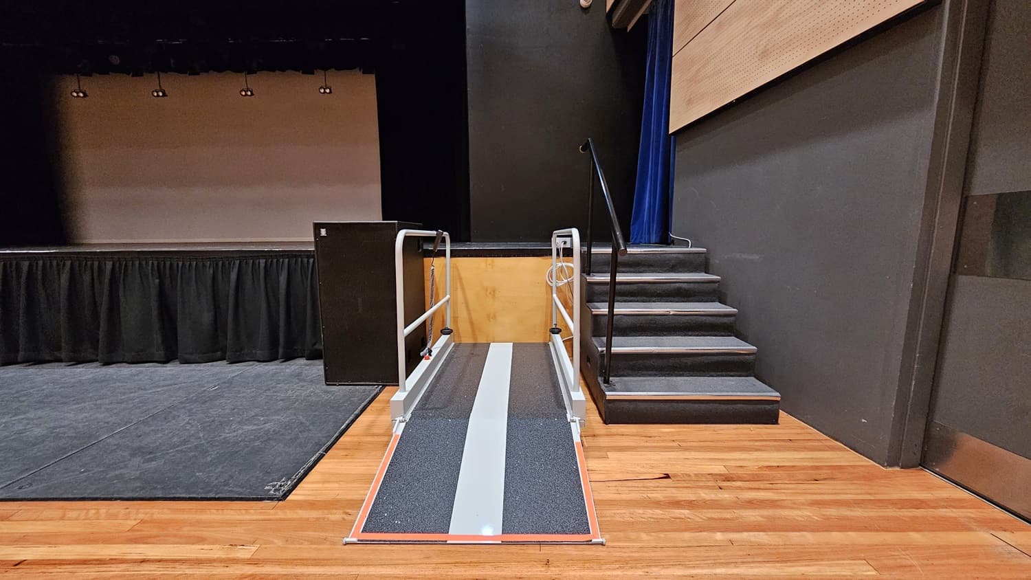 On the left, there is a stage with black curtains and a black floor. On the right, there is a set of black stairs with a handrail leading up to the stage. In the middle, there is a grey ramp with handrails on both sides, leading up to the stage. The background features a beige wall with black curtains above. The floor is polished light wood, and the ceiling is black with recessed lights.