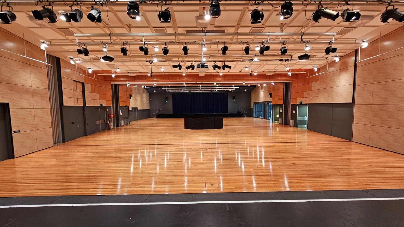 On the left, there are beige wooden walls with black doors and a fire extinguisher mounted on the wall. On the right, there are similar beige wooden walls with black doors and a green exit sign above one door. In the middle, there is a large open space with polished light wood flooring and a black stage with blue curtains in the background. The ceiling is white with a grid pattern, featuring numerous recessed lights and black stage lighting equipment.