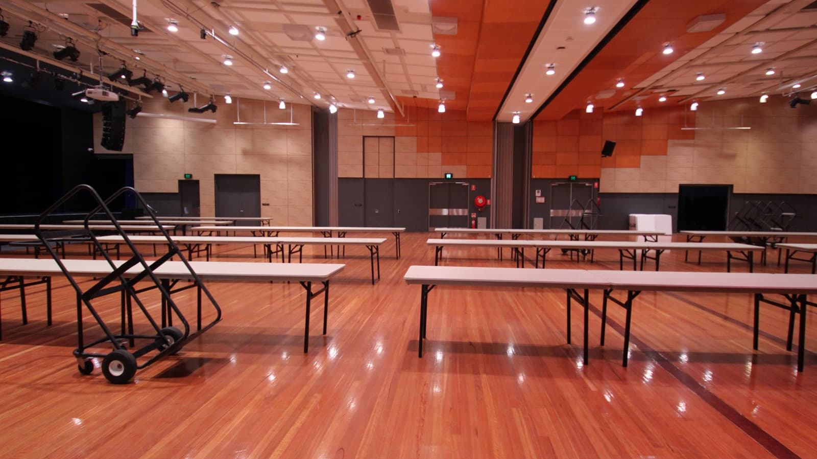 On the left, there are black double doors and a stage area with black curtains and lighting equipment. On the right, there are beige walls with black doors and a green exit sign above one door. In the middle, there are several long tables arranged in rows, with a black trolley holding folded tables. The background features beige and orange acoustic panels on the walls. The floor is polished light wood, and the ceiling is white with a grid pattern and numerous recessed lights.