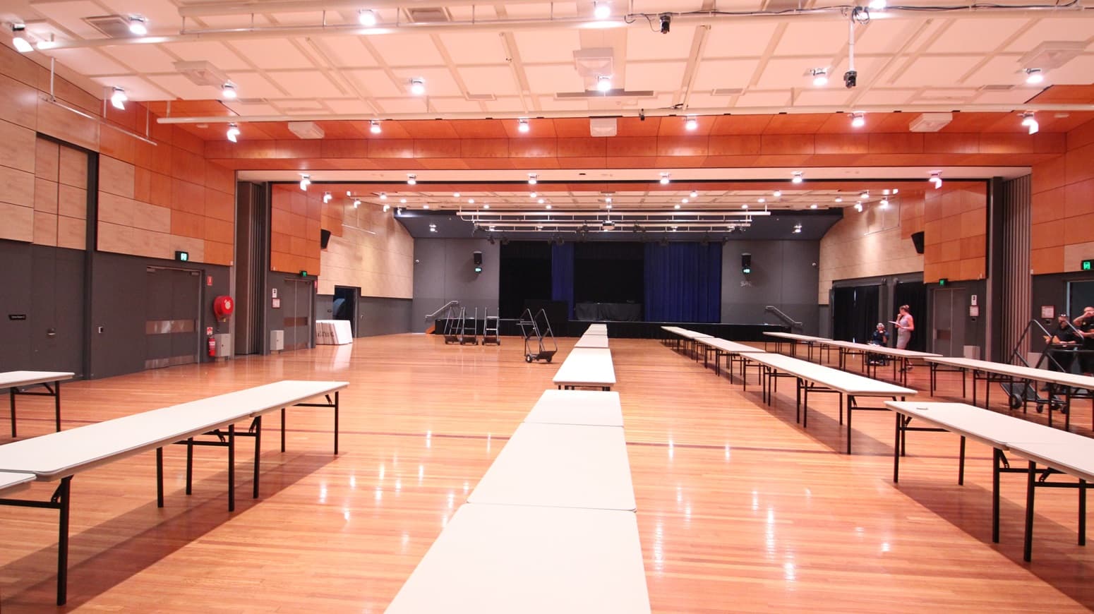 On the left, there are beige wooden walls with black double doors and a fire extinguisher mounted on the wall. On the right, there are similar beige wooden walls with black doors and a green exit sign above one door. In the middle, there are long white tables arranged in rows on a polished light wood floor. The background features a stage with blue curtains and black skirting. The ceiling is white with a grid pattern, recessed lights, and black stage lighting equipment.
