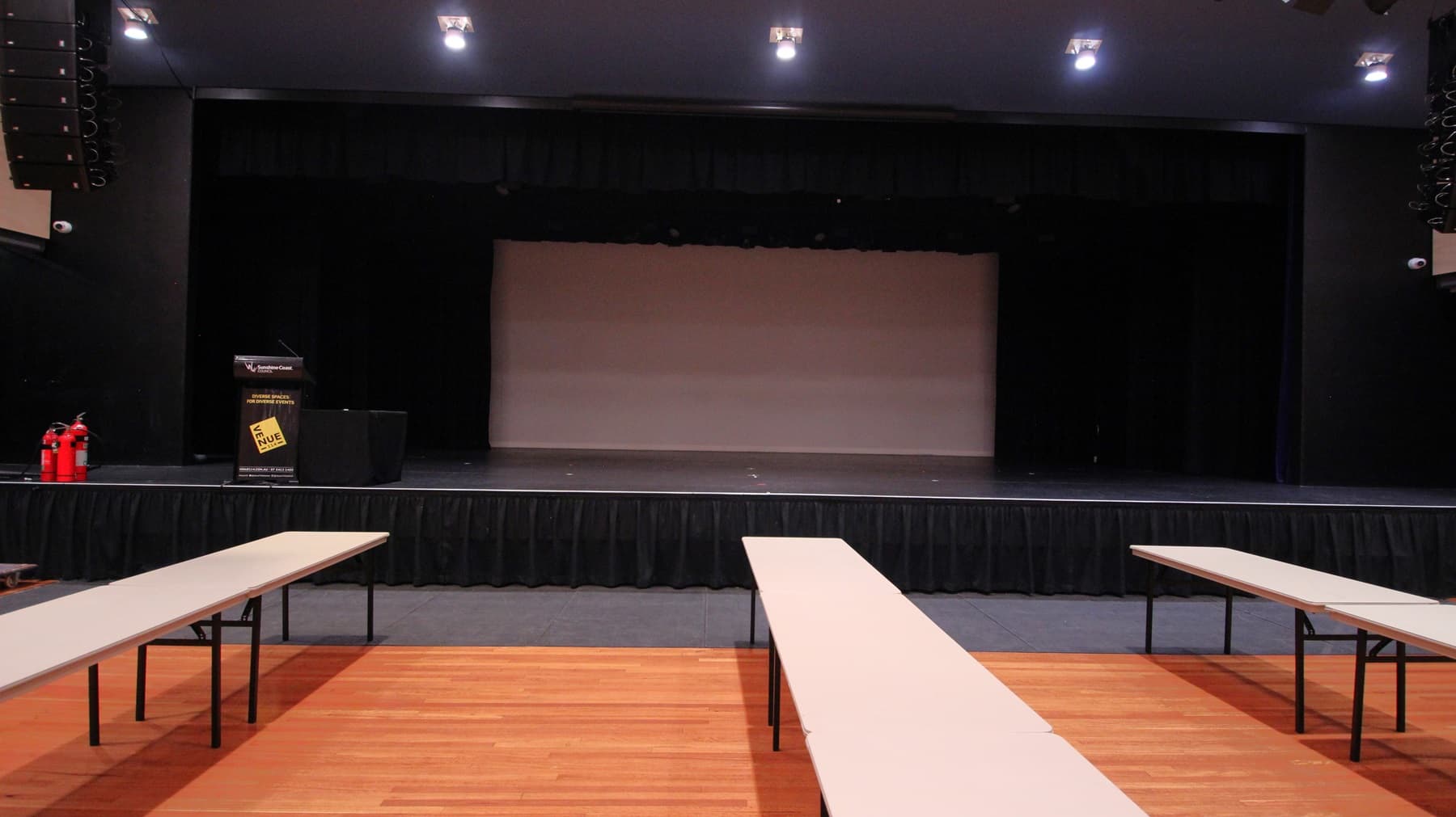 On the left, there are two red fire extinguishers on the floor next to a black podium with a sign. On the right, there is a black wall with stage lighting equipment. In the middle, there is a large stage with black curtains and a grey backdrop. The foreground features long white tables arranged in rows on a polished light wood floor. The ceiling is dark with recessed lights and additional stage lighting equipment.
