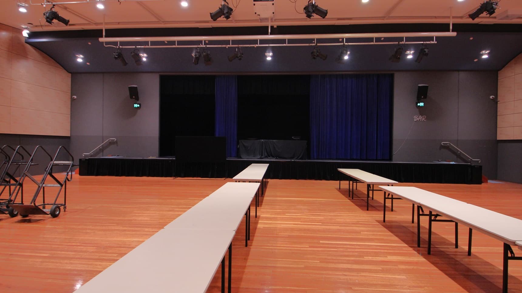 On the left, there are beige walls with black lower sections and two trolleys holding folded tables. On the right, there are similar beige walls with black lower sections and a green exit sign above a door. In the middle, there are long white tables arranged in rows on a polished light wood floor. The background features a stage with blue curtains and black skirting. The ceiling is white with recessed lights and black stage lighting equipment.