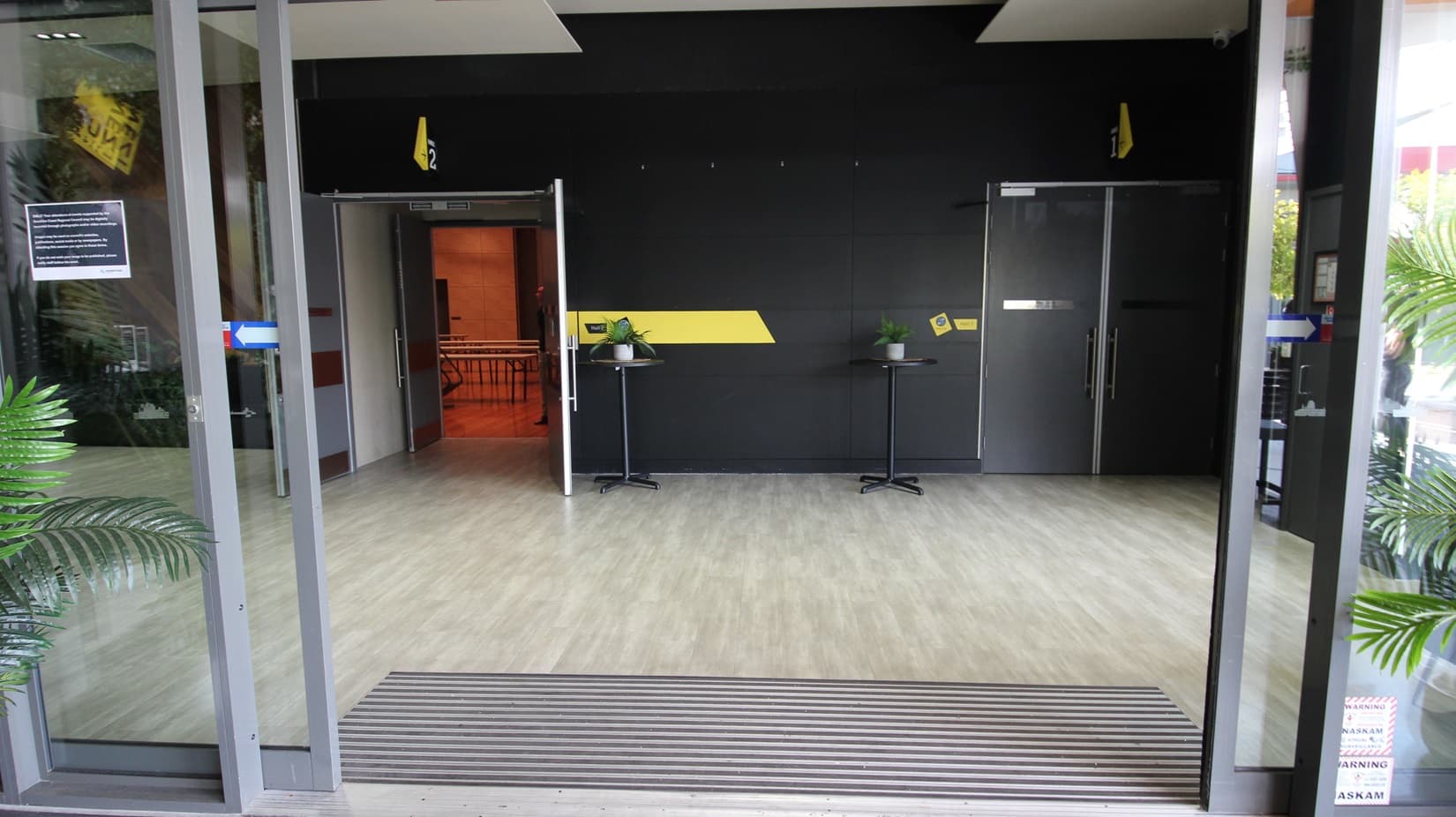 On the left, there is a glass door with a potted plant next to it. On the right, there is another glass door with a potted plant beside it. In the middle, there is a black wall with a yellow diagonal stripe, two small round tables with plants on them, and two grey doors. The floor is light wood, and the ceiling is black with recessed lights.