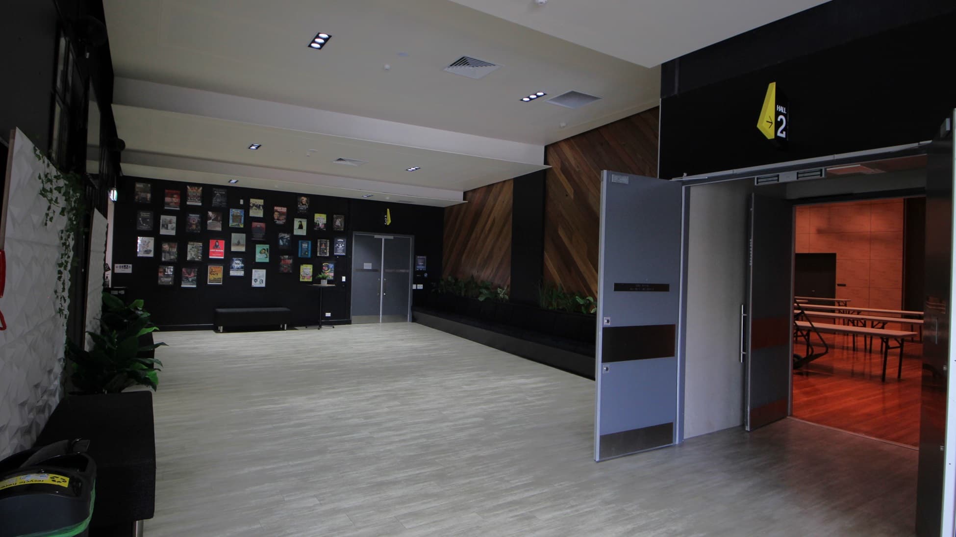 On the left, there is a black bench with potted plants and a wall covered with framed pictures. On the right, there are two open grey doors leading to a room with wooden flooring and tables. In the middle, there is an open space with light wood flooring. The background features a black wall with a yellow sign and a section of wooden panelling. The ceiling is white with recessed lights and ventilation panels.
