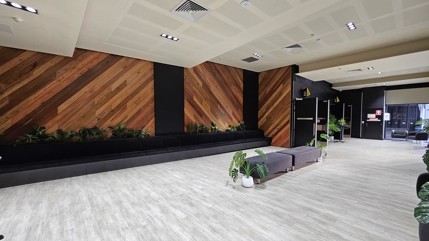 On the left, there is a black bench with potted plants and a wall covered with framed pictures. On the right, there are two open grey doors leading to a hallway with light wood flooring and more framed pictures. In the middle, there are two black armchairs with a small table and potted plants. The background features a black wall with a whiteboard and additional framed pictures. The ceiling is white with recessed lights and ventilation panels. The floor is light grey wood.