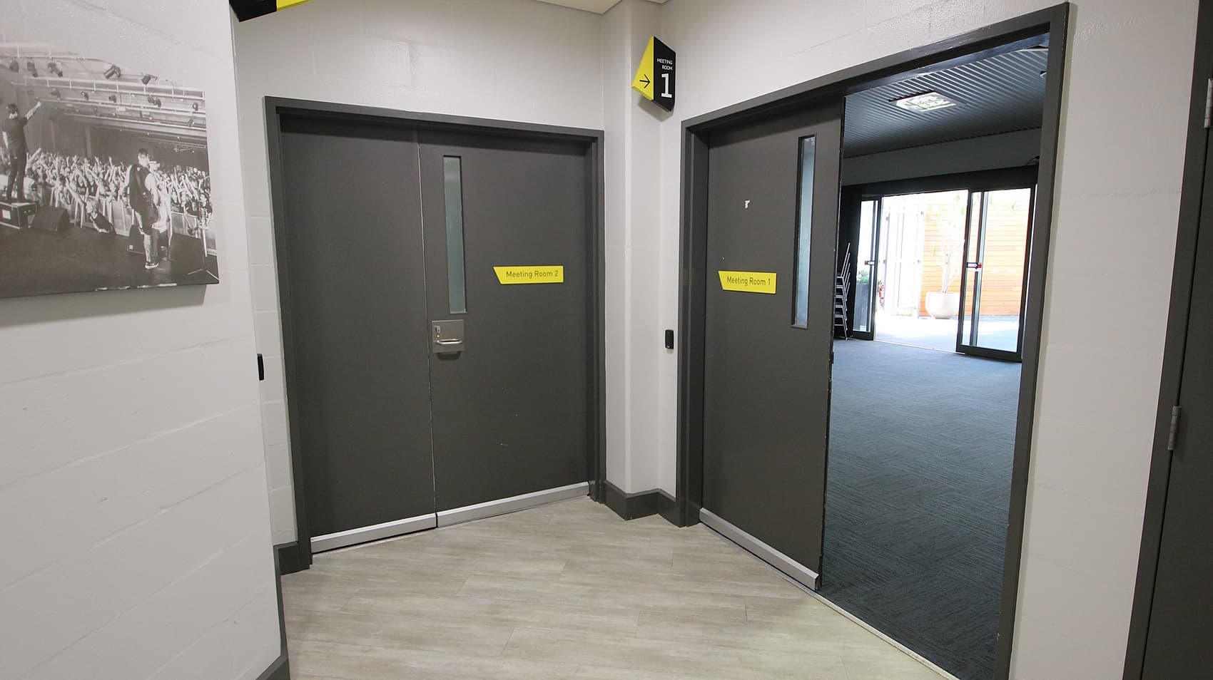 On the left, there is a black and white photograph of a crowd on a light grey wall. Below the photograph, there are two grey double doors with yellow signs above them. On the right, there is an open doorway leading to a room with dark carpet and large windows. The floor is light grey wood, and the ceiling is grey with recessed lights and ventilation panels.