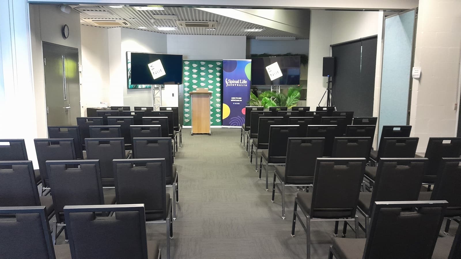 On the left, there is a wall with a closed door and a mounted television screen. On the right, there is a wall with a window covered by a black roller blind and a potted plant in front of it. In the middle, there are rows of black chairs facing a wooden podium with a microphone, flanked by two banners and another television screen. The floor is covered in dark grey carpet, and the ceiling is white with recessed lights and ventilation panels.