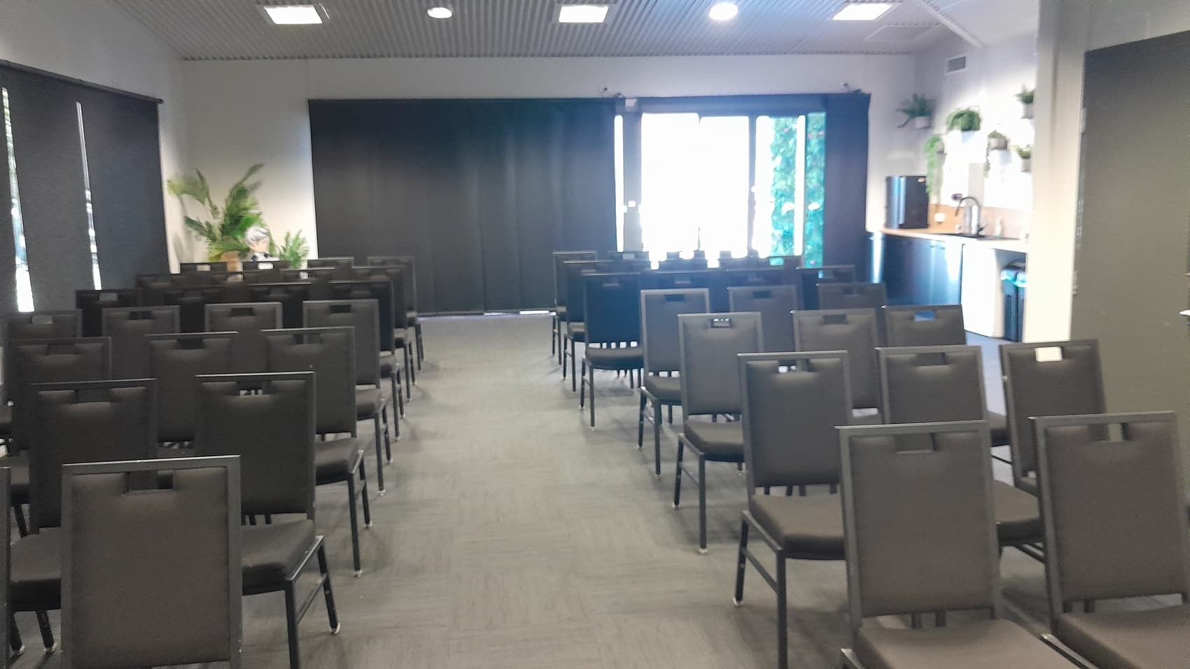 On the left, there are rows of black chairs facing forward, with a wall featuring black roller blinds and potted plants. On the right, there are more rows of black chairs, a countertop with a coffee machine, cups, and potted plants above. In the background, there is a large window with black curtains partially open, revealing greenery outside. The floor is covered in dark grey carpet, and the ceiling is white with recessed lights and ventilation panels.
