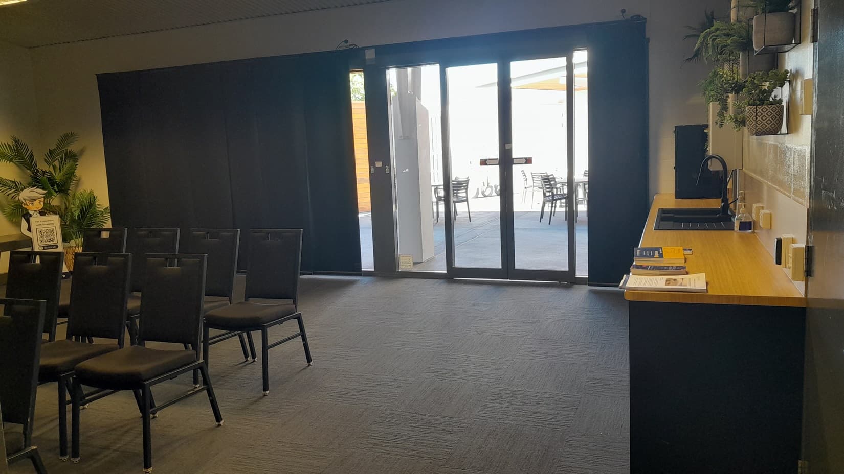 On the left, there are rows of black chairs facing forward, with a potted plant in the corner. On the right, there is a countertop with a coffee machine, cups, books, and potted plants on shelves above. In the background, there are large glass doors leading to an outdoor area with tables and chairs. The floor is covered in dark grey carpet tiles, and the ceiling is white with recessed lights.