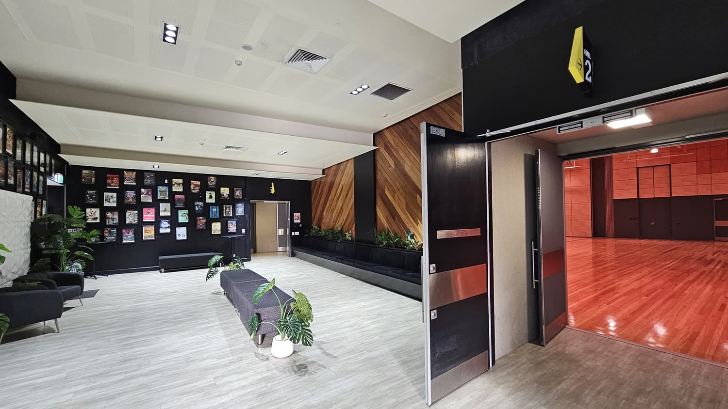 On the left, there is a black bench with potted plants and a wall covered with framed pictures. On the right, there are two open grey doors leading to a room with a wooden floor and red walls. In the middle, there are two black armchairs with a small table and potted plants. The background features a black wall with diagonal wooden panels and additional potted plants. The floor is light grey wood, and the ceiling is white with recessed lights and ventilation panels.