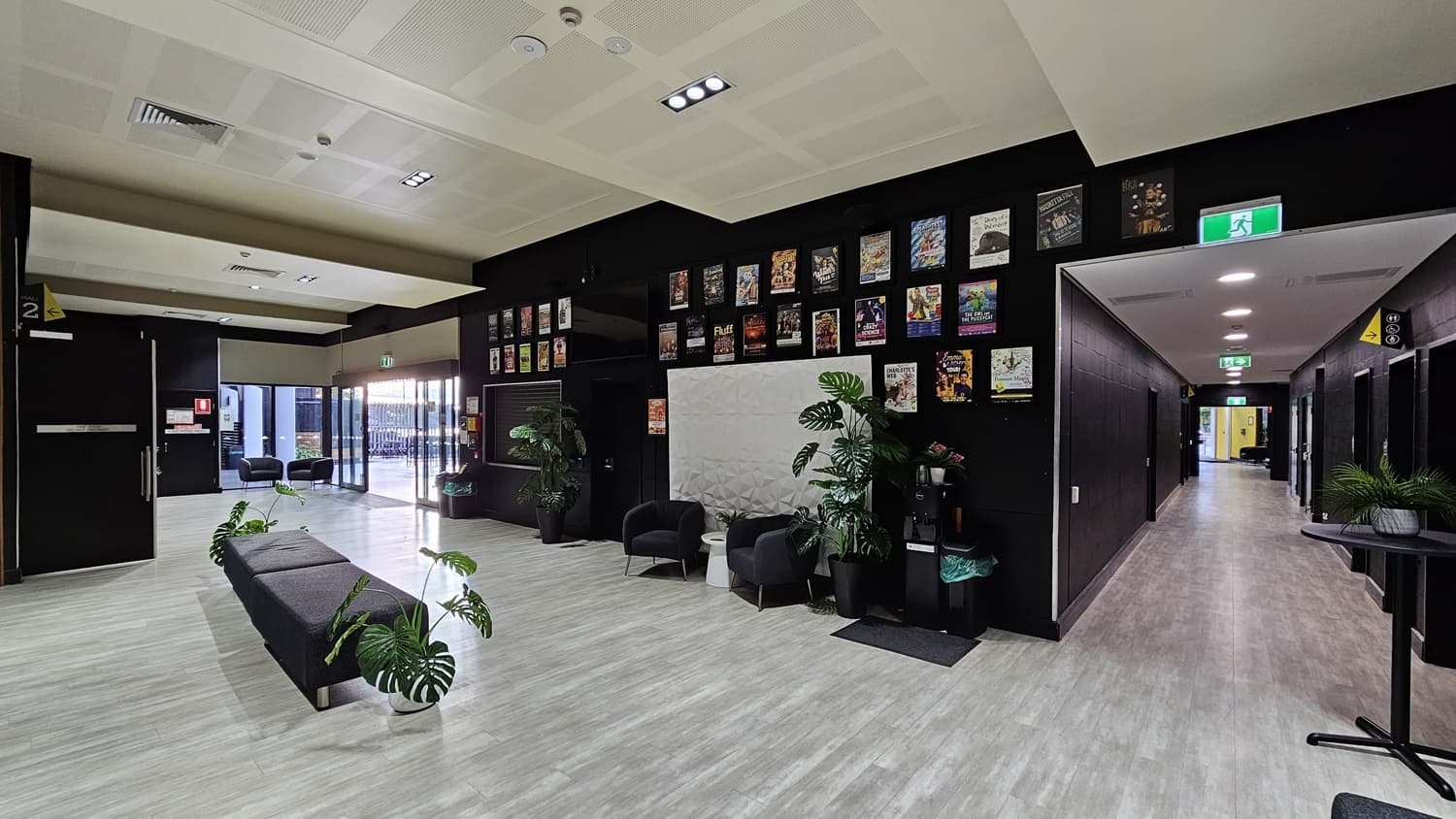 On the left, there is a long black bench with potted plants on the floor and a wall with framed posters above. On the right, there is a corridor with a light grey floor leading to a brightly lit area, and a black wall with a whiteboard and two black armchairs with potted plants. In the background, there are glass doors opening to an outdoor area with more seating. The floor is light grey wood, and the ceiling is white with recessed lights and ventilation panels.