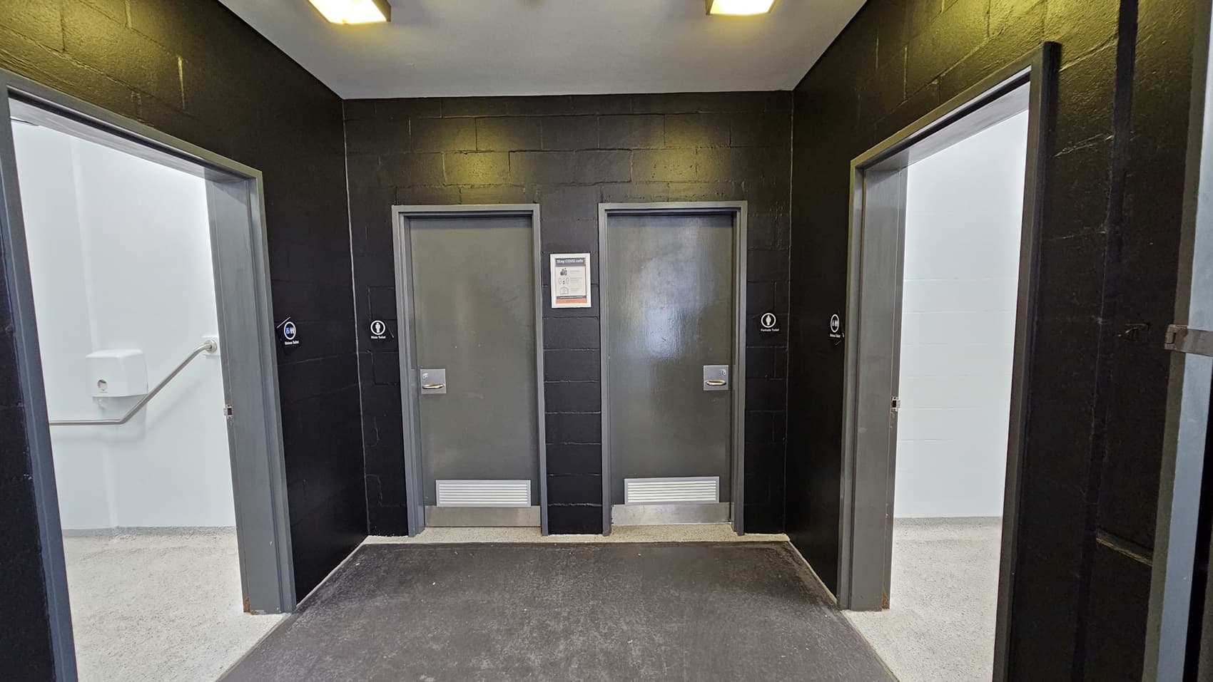 On the left, there is a grey door with a toilet sign and a white wall with a handrail. On the right, there is another grey door with a toilet sign and a white wall. In the middle, there are two grey doors with a framed notice between them. The floor is a mix of dark grey near the doors and light grey in the foreground. The ceiling is white with recessed rectangular lights.