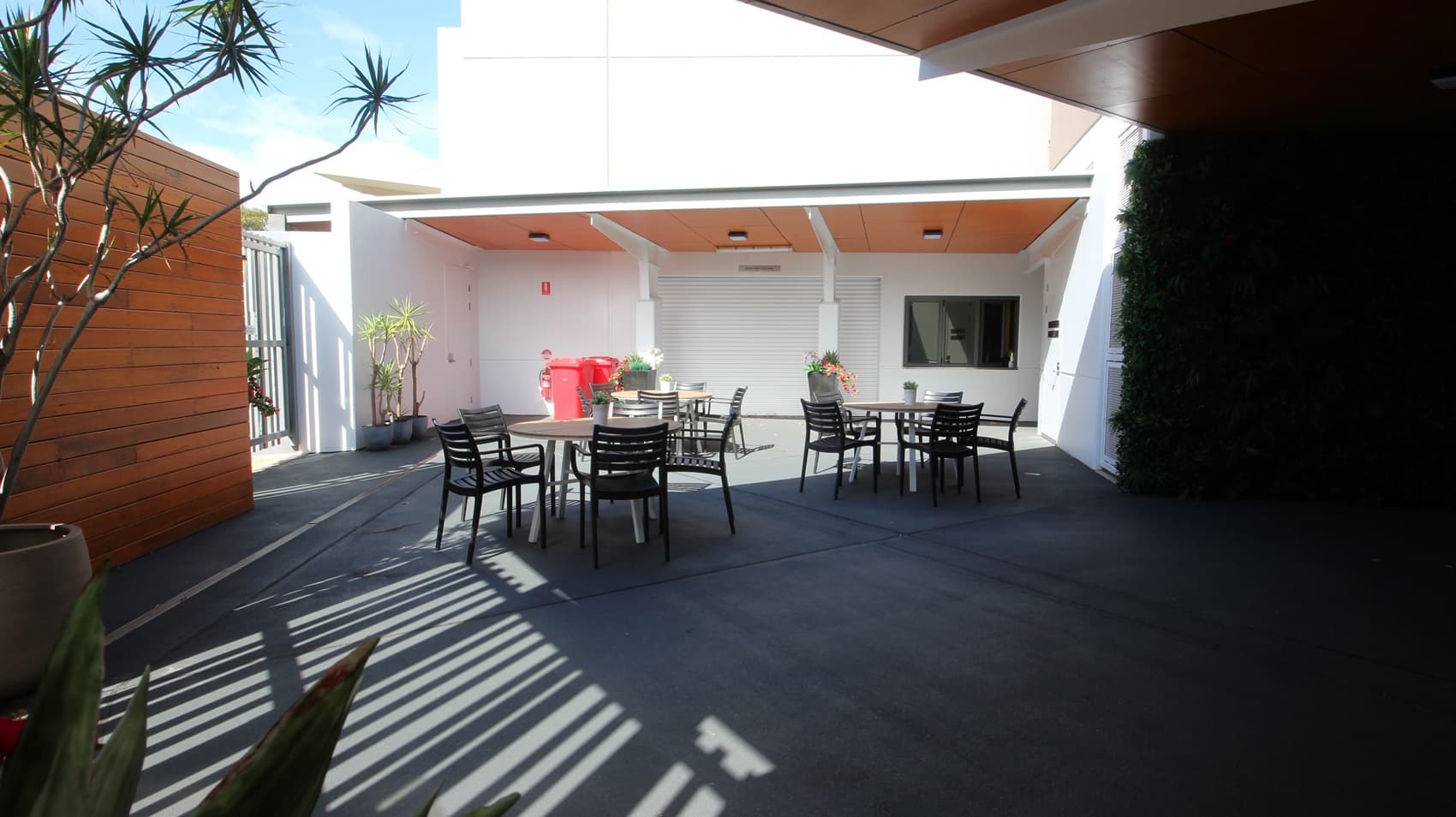 On the left, there is a wooden wall with a tall potted plant in front of it. On the right, there is a dark wall covered with greenery. In the middle, there are several black tables with black chairs arranged around them. In the background, there is a white building with a wooden ceiling overhang, a red fire extinguisher, and potted plants near a closed white roller door. The floor is dark grey concrete, and the ceiling under the overhang is wooden with recessed lights.