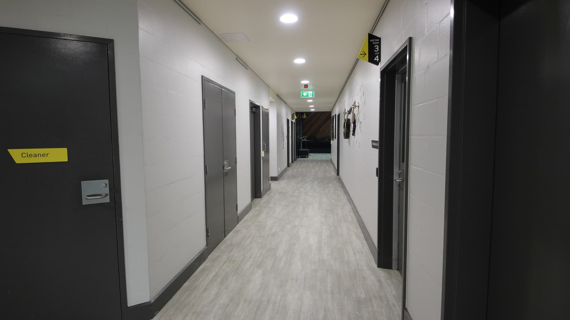 On the left, there are grey doors with yellow signs, including one marked "Cleaner." On the right, there are more grey doors, one with a sign and hooks holding cleaning equipment. In the middle, a long corridor with a light grey wood-effect floor leads to a distant area with a green exit sign above a door. The ceiling is white with recessed lights and ventilation panels.