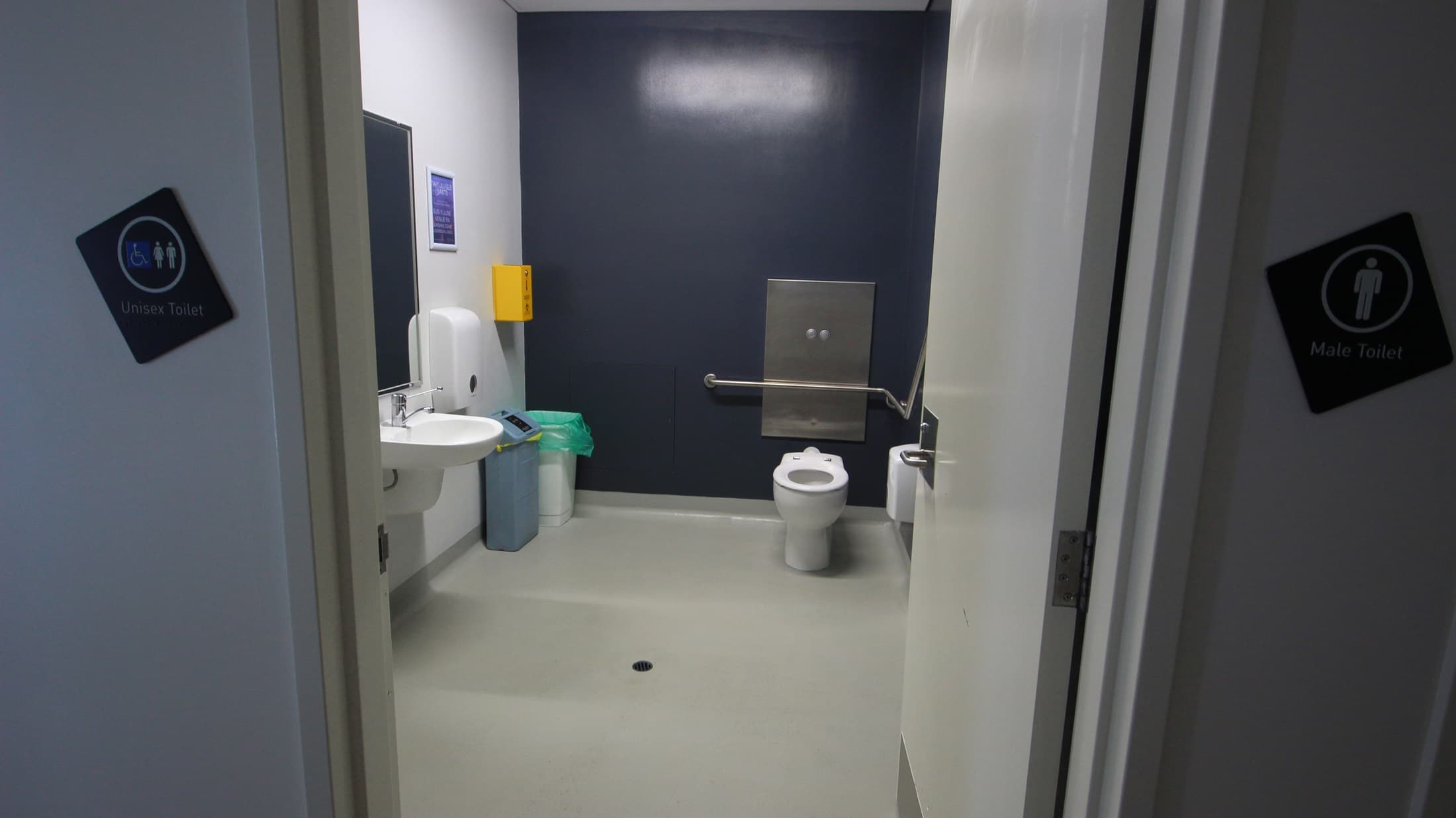 On the left, there is a white sink with a soap dispenser above it, a paper towel dispenser, and a green bin below. On the right, there is a white wall with a handrail extending from the back wall. In the middle, there is a white toilet with a metal flush panel above it and a yellow bin to the left. The floor is light grey, and the ceiling is white with no visible fixtures. The walls are a mix of white and dark blue.