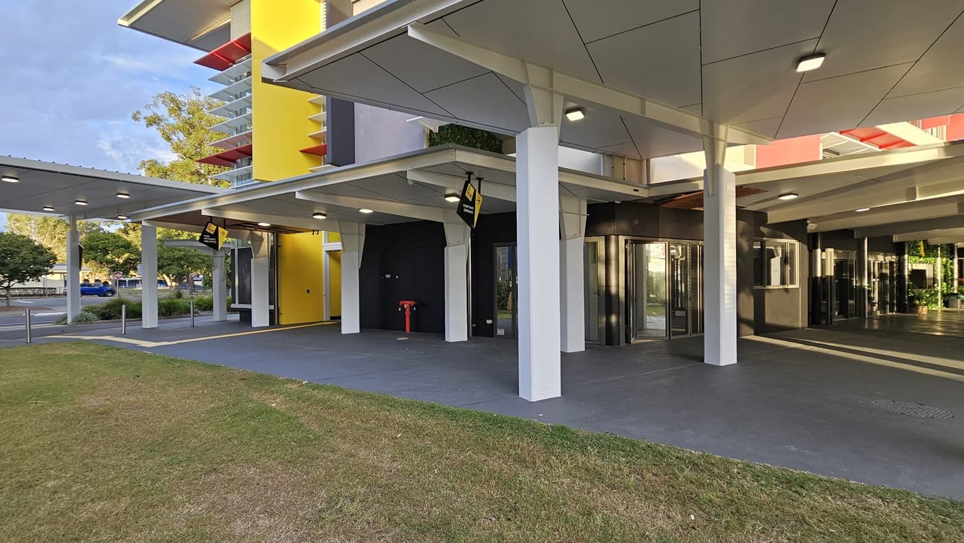 On the left, there is a yellow wall with a door and a black wall with a yellow sign. On the right, there are glass revolving doors leading into a building with a black wall. In the middle, there are white pillars supporting a white ceiling with recessed lights. The floor is grey concrete, and the foreground has a grassy area. The background includes additional white pillars and a yellow vertical structure with balconies.