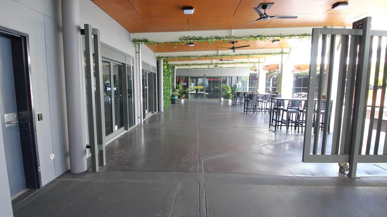 On the left, there are grey walls with glass doors and windows, and a black door. On the right, there are tall metal gates and a seating area with high tables and chairs. In the middle, there is a wide walkway with a dark concrete floor leading to more glass doors and potted plants. The ceiling is wooden with recessed lights and ceiling fans, and it has green vines hanging from it.