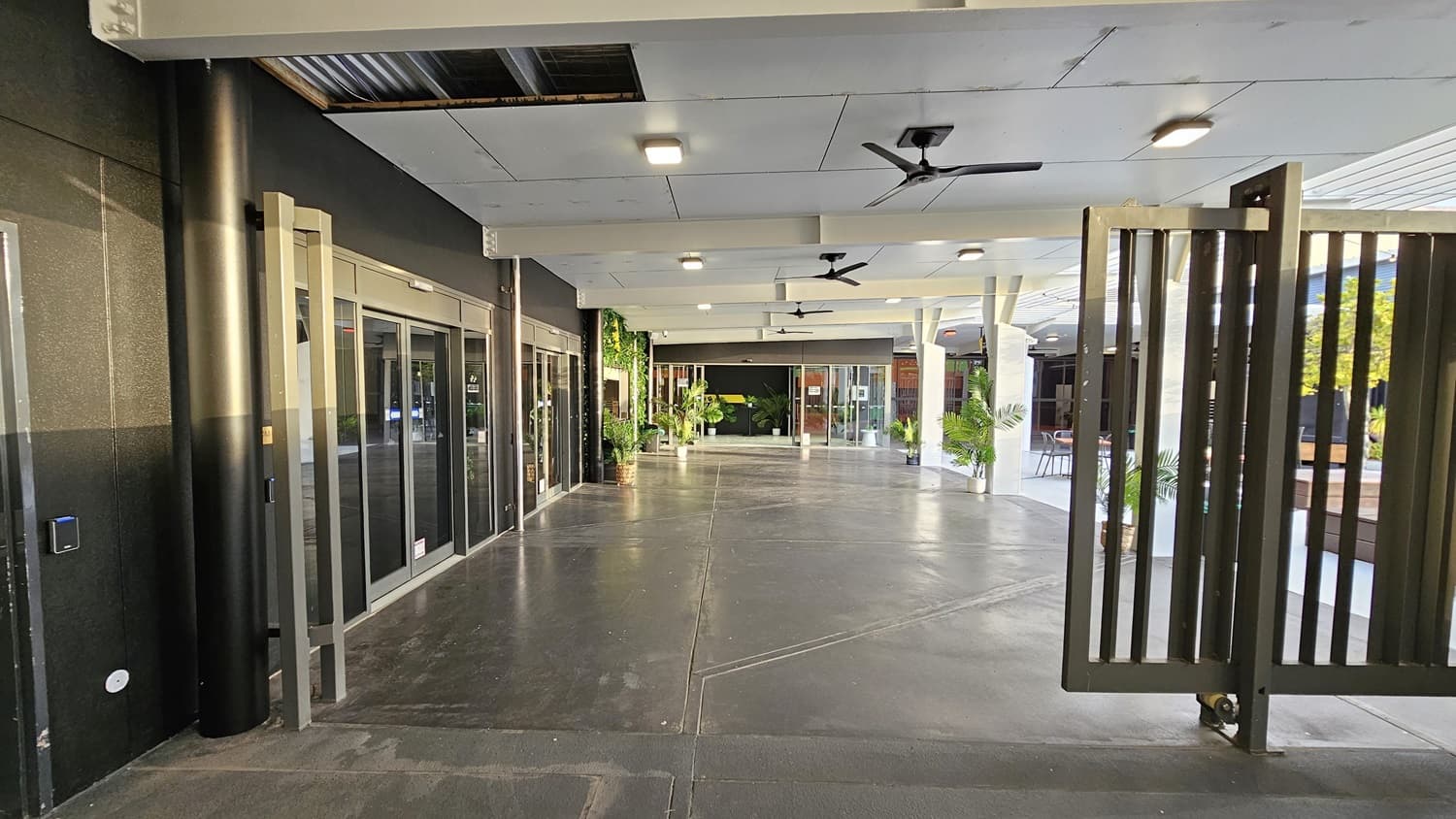 On the left, there are black walls with large glass windows and doors, and a black pillar. On the right, there are tall metal gates and a seating area with tables, chairs, and potted plants. In the middle, there is a wide walkway with a dark concrete floor leading to more glass doors and greenery. The ceiling is white with recessed lights and ceiling fans.