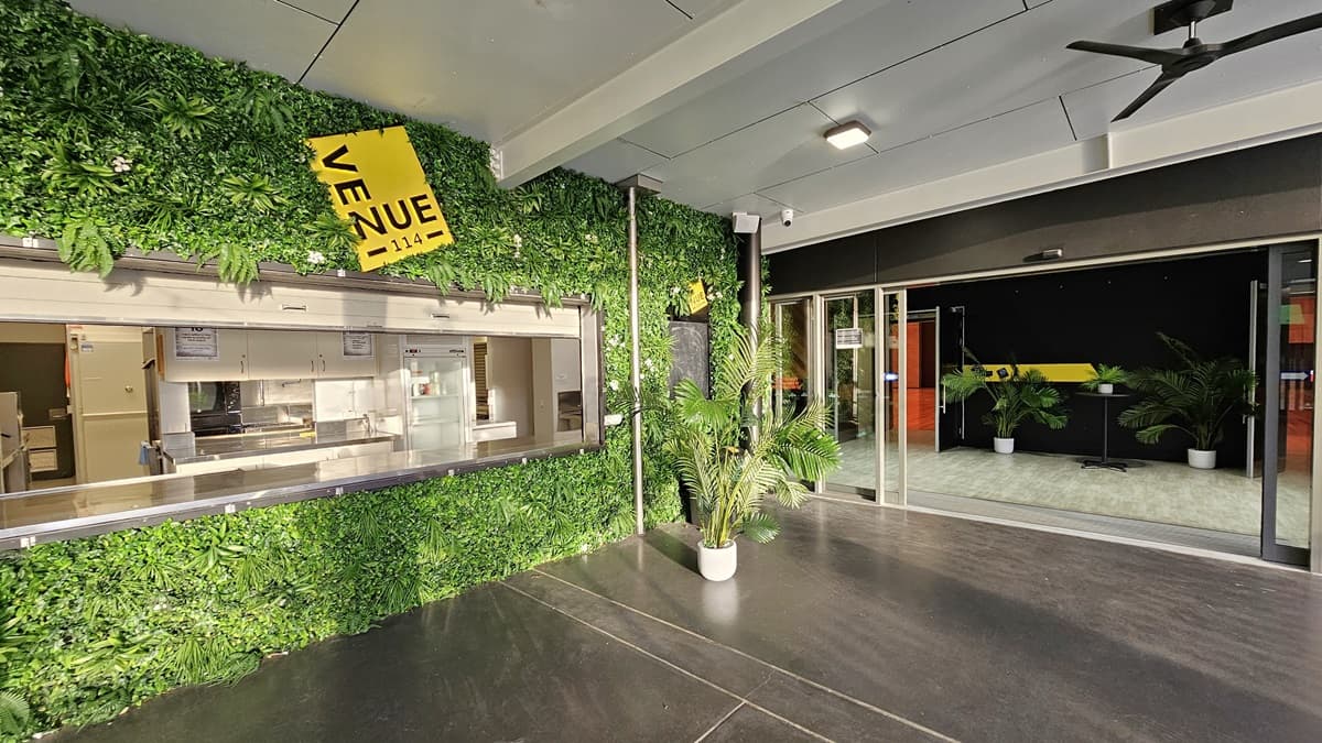 On the left, there is a green wall covered with artificial foliage and a yellow sign that reads "VENUE." Below the sign, there is a serving window with a kitchen visible inside. On the right, there are glass sliding doors leading to an indoor area with a black and yellow wall, potted plants, and a light-coloured floor. In the middle, there is a dark concrete floor. The ceiling is white with recessed lights and a ceiling fan.