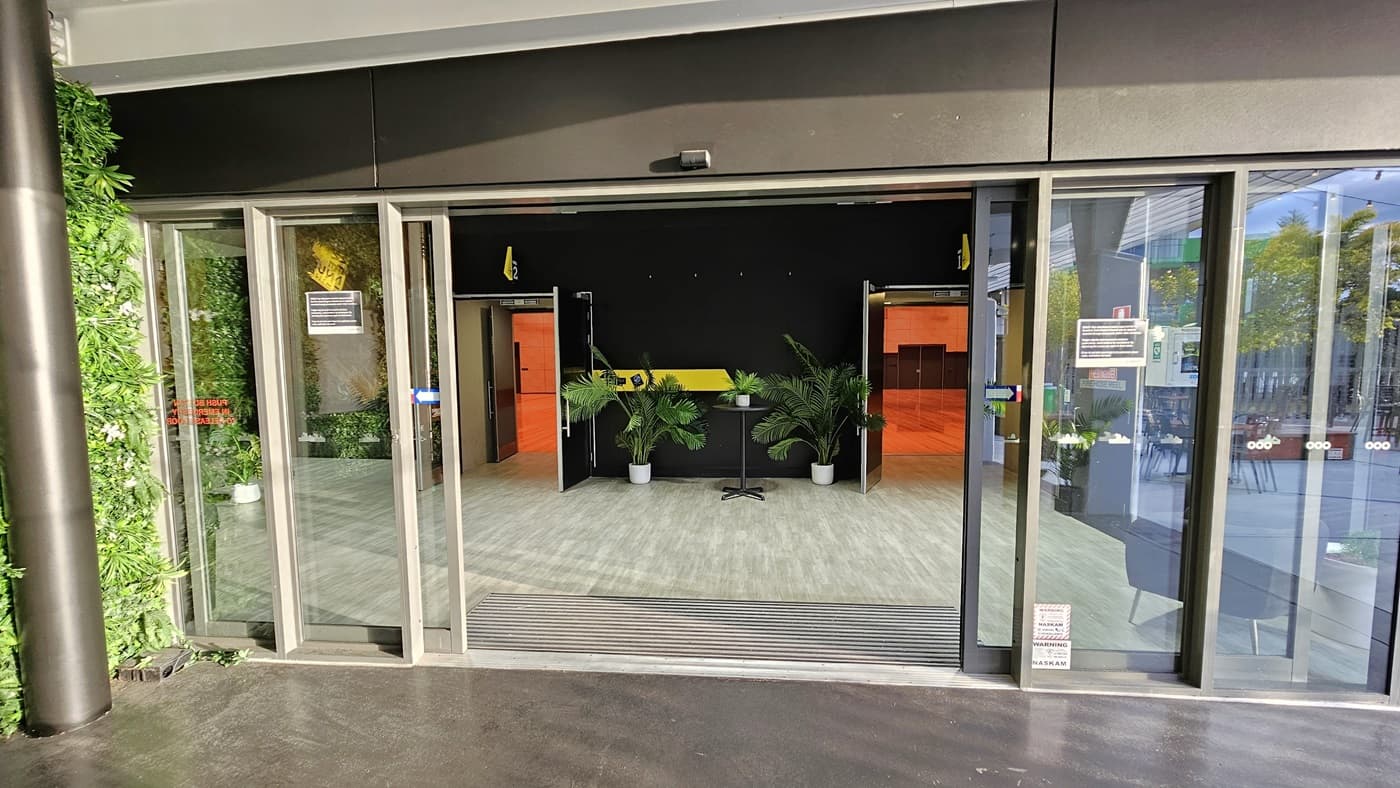 On the left, there is a green wall covered with artificial foliage. On the right, there are glass sliding doors leading to an outdoor seating area with tables and chairs. In the middle, there is an entrance with glass sliding doors opening into a room with a light wood-effect tile floor, black walls, and potted plants. The ceiling inside is white with recessed lights.