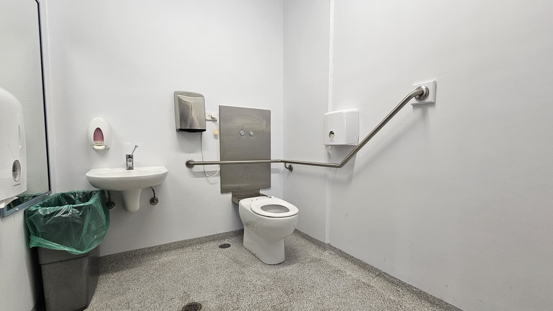 On the left, there is a white sink with a soap dispenser above it, a paper towel dispenser, and a green bin below. On the right, there is a white toilet with a metal grab bar extending along the wall. In the middle, there is a grey panel behind the toilet with two flush buttons. The floor is speckled grey, and the walls and ceiling are white.