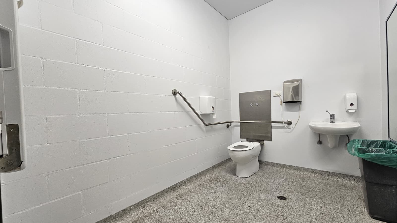 On the left, there is a white wall with a metal grab bar extending horizontally. On the right, there is a white sink with a soap dispenser above it, a paper towel dispenser, and a green bin below. In the middle, there is a white toilet with a metal grab bar on the wall behind it and a grey panel with two flush buttons. The floor is speckled grey, and the ceiling is white.