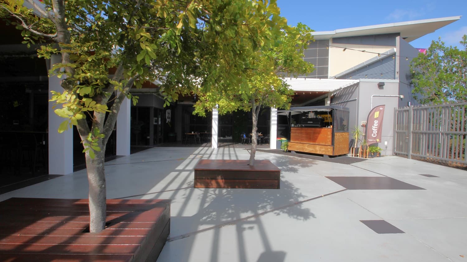 On the left, there are two trees planted in wooden benches. On the right, there is a wooden kiosk with a "Coffee" banner next to it and a grey fence behind. In the middle, there is an open concrete area with shadows of the trees. The background features a building with large windows, a white overhang, and a grey upper section. The floor is light grey concrete, and the ceiling of the kiosk area is white with recessed lights.