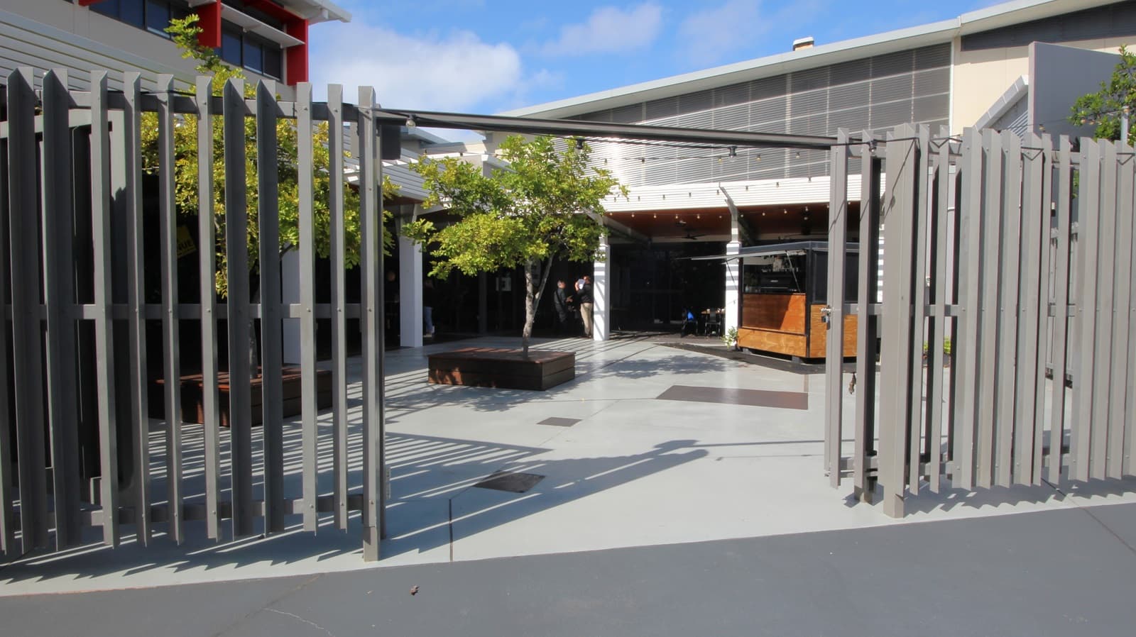 On the left, there is a grey metal fence with vertical slats. On the right, there is a continuation of the same grey metal fence. In the middle, there is an open courtyard with light grey concrete flooring, a wooden bench surrounding a tree, and a person standing near the centre. The background features a building with large windows, a white overhang, and a grey upper section with horizontal slats.