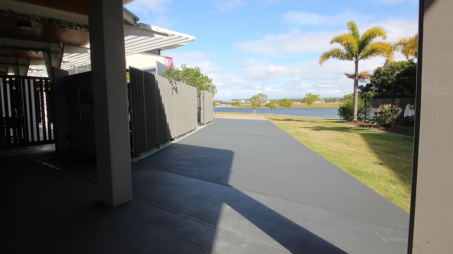 On the left, there is a covered area with a grey metal fence and a white overhang. On the right, there is a grassy lawn with a palm tree and some shrubs. In the middle, there is a wide grey concrete pathway leading towards a body of water in the background. The floor under the covered area is dark grey concrete, and the ceiling is white with recessed lights.