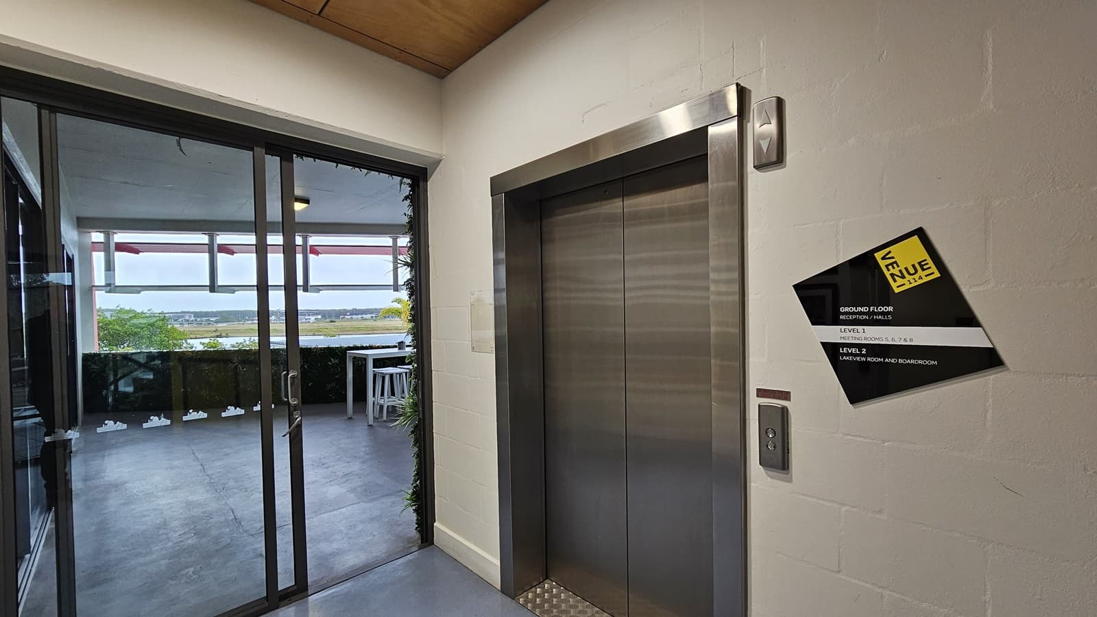 On the left, there is a glass sliding door leading to a balcony with a view of a distant landscape. The balcony has a white table and chairs. On the right, there is a metallic lift with a control panel and a black and yellow sign indicating floor information. In the middle, a white wall separates the lift from the glass door. The floor is grey and appears to be concrete, while the ceiling is wooden.