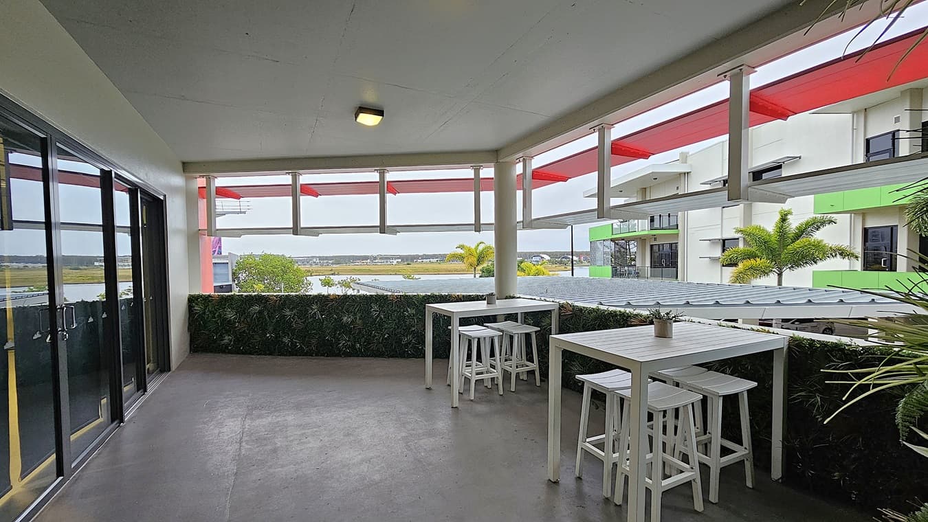 On the left, there are glass sliding doors with black frames. On the right, there are white railings and a view of adjacent buildings with green and white exteriors. In the middle, there are two white high tables with matching stools, and a small plant on one of the tables. The background features a distant landscape with trees and buildings. The floor is grey and appears to be concrete, while the ceiling is white with a single light fixture.