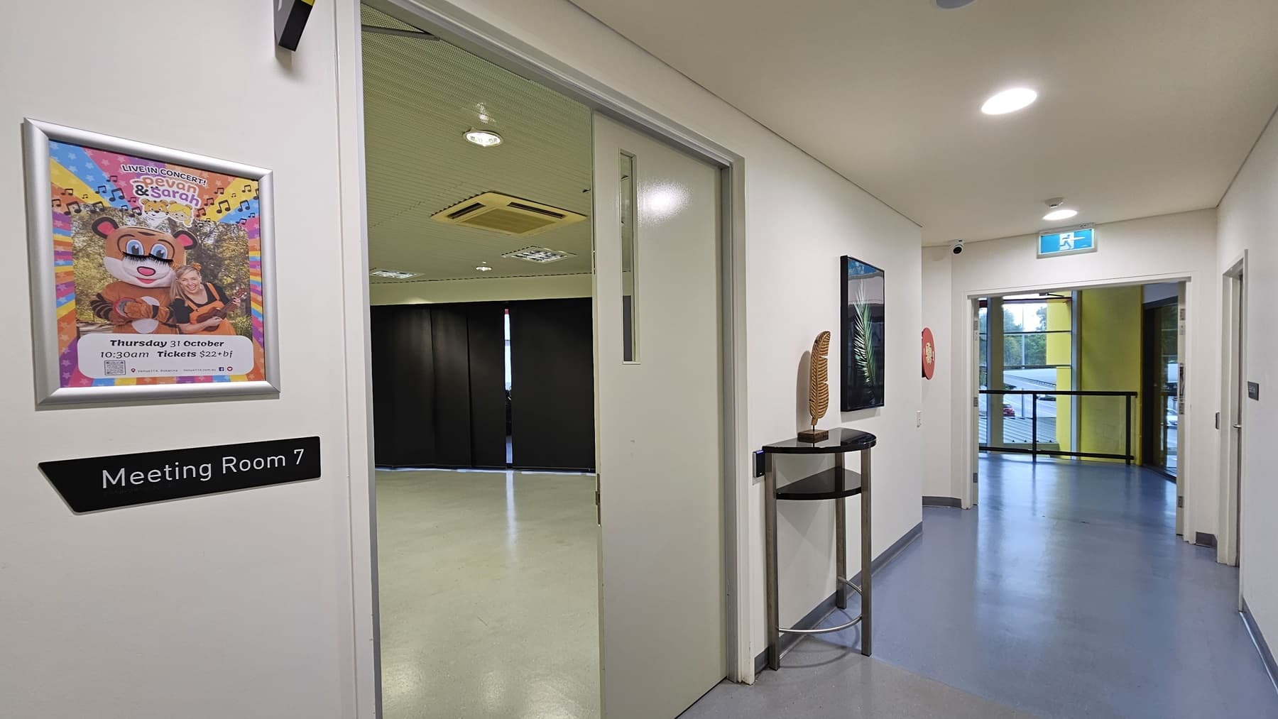 On the left, there is an open door with a sign reading "Meeting Room 7" and a colourful poster above it. Inside the room, there are black curtains covering the windows and a beige floor. On the right, there is a corridor with a fire extinguisher mounted on the wall and a small table with a decorative item. The background features a glass door leading to a balcony with yellow walls. The floor is light grey, and the ceiling is white with recessed lighting and an air conditioning unit.