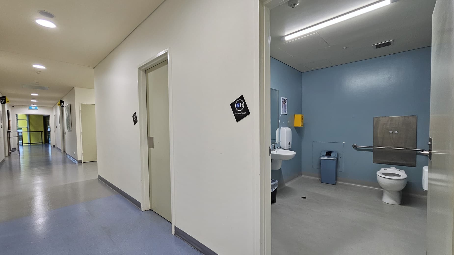 On the left, there is a long corridor with several closed doors, each with a small black sign. The corridor has a light blue and grey floor and white walls. On the right, there is an open door leading to a bathroom with light blue walls, a white toilet, a sink, a soap dispenser, and a blue bin. In the background, the corridor continues with more doors and a bright light at the end. The ceiling is white with recessed lighting and a ventilation unit.
