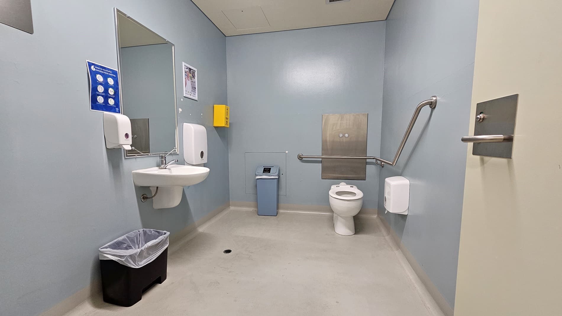 On the left, there is a white sink with a mirror above it, a soap dispenser, and a paper towel dispenser. Below the sink, there is a black bin with a liner. On the right, there is a white toilet with a grab rail on the wall, a toilet paper dispenser, and a small blue bin next to it. In the middle, there is a yellow box mounted on the wall. The floor is light grey, and the ceiling is white with a ventilation panel.